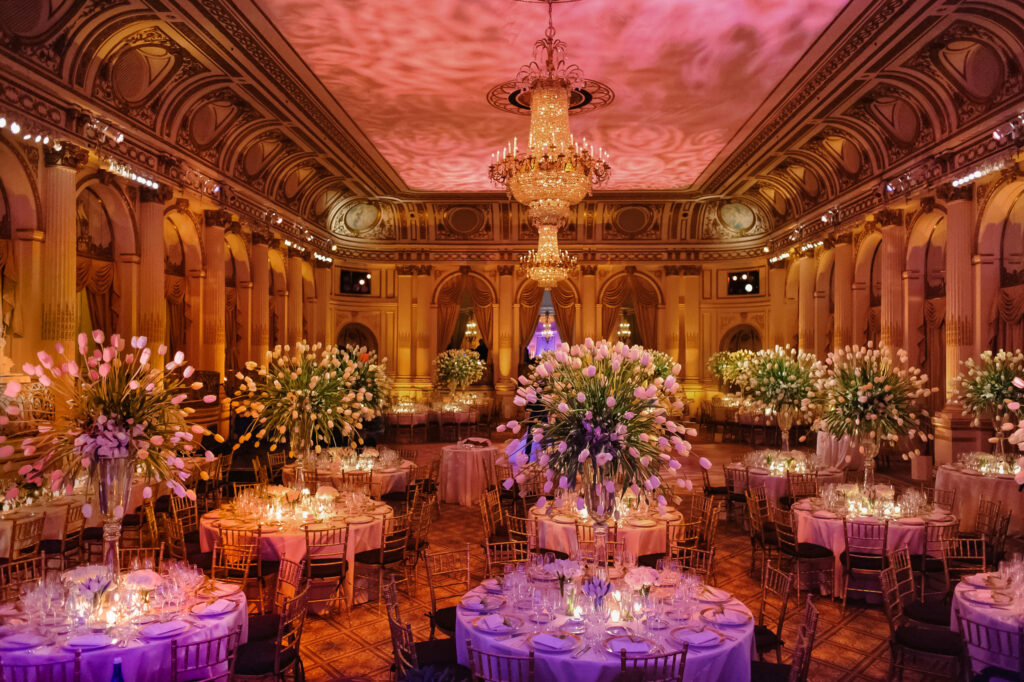 A grand ballroom features ornate chandeliers, tall floral centerpieces, elegantly set round tables, and warm ambient lighting, perfect for those learning how to plan a wedding weekend in a luxurious event space.