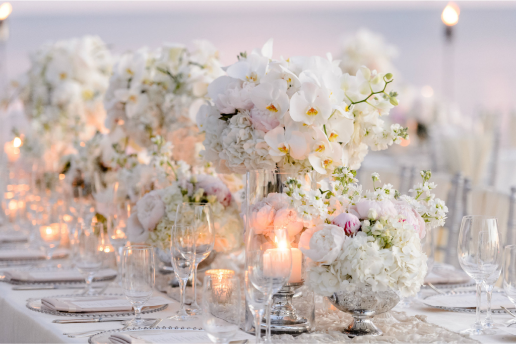 A long, elegantly set table features white floral arrangements, clear glassware, and glowing candles, creating a sophisticated dining ambiance perfect for a wedding weekend.