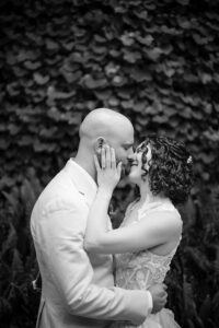 The bride and groom share an intimate moment, touching foreheads and smiling with closed eyes. The groom, dressed in a light suit, gently embraces the bride, who is wearing a lace gown. The lush, leafy background enhances the romantic and serene ambiance of the photo.