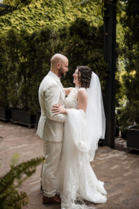 The bride and groom share a joyful embrace outdoors, surrounded by lush greenery. The groom wears a light suit, and the bride is dressed in a lace gown with a long veil. They gaze into each other's eyes, smiling, capturing a tender moment of happiness on their wedding day.