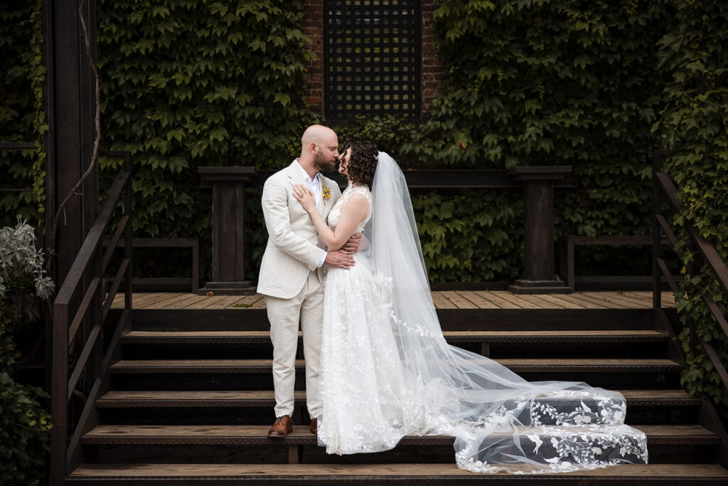 A bride and groom share a kiss on a set of wooden steps surrounded by lush green ivy. The groom is dressed in a light-colored suit, while the bride wears a detailed lace wedding gown with a long veil cascading down the steps. The intimate moment is framed by rustic wooden railings and a serene, natural backdrop.