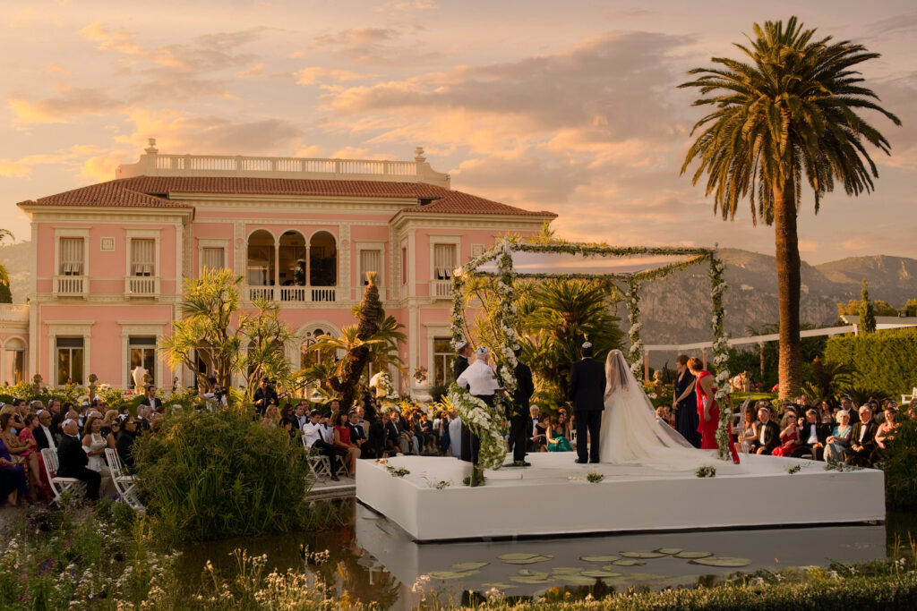 A couuple stands in front of their family and friends during a wedding ceremony under a pink orange sky.