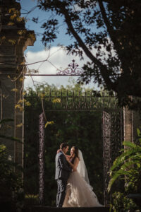 A couple hugs one another, surrounded by arches and greenery.