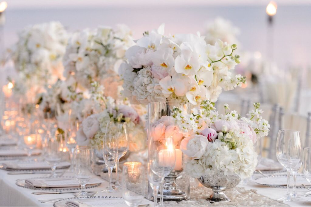 A long, elegantly set table features white floral arrangements, clear glassware, and glowing candles