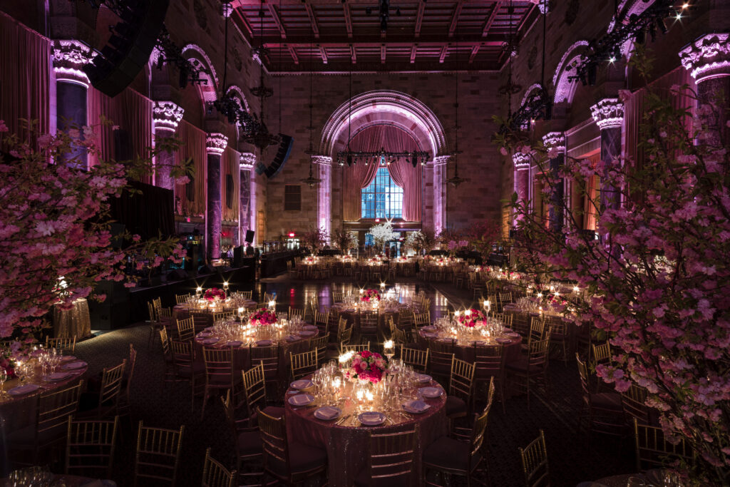 A grand, dimly lit ballroom at Gotham Hall is set for an event with round tables adorned with floral centerpieces and candles. The room, often listed among the best indoor wedding venues in NYC, is decorated with pink lighting and towering floral arrangements.