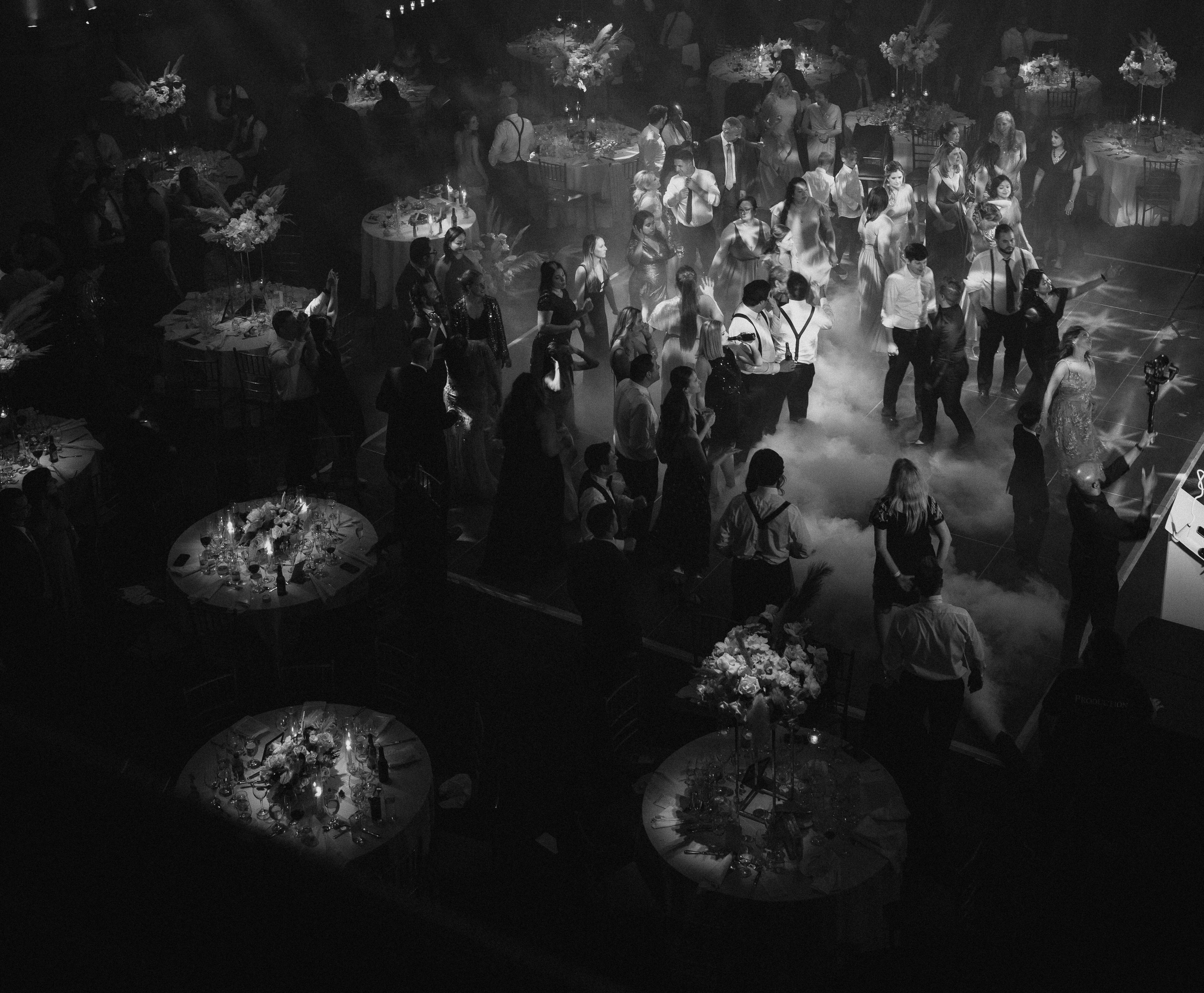 A black and white photograph by NYC Photography for Destination Weddings shows a crowd of people dancing and mingling at a formal event with round tables and floral centerpieces. Smoke or fog effects are present on the dance floor.