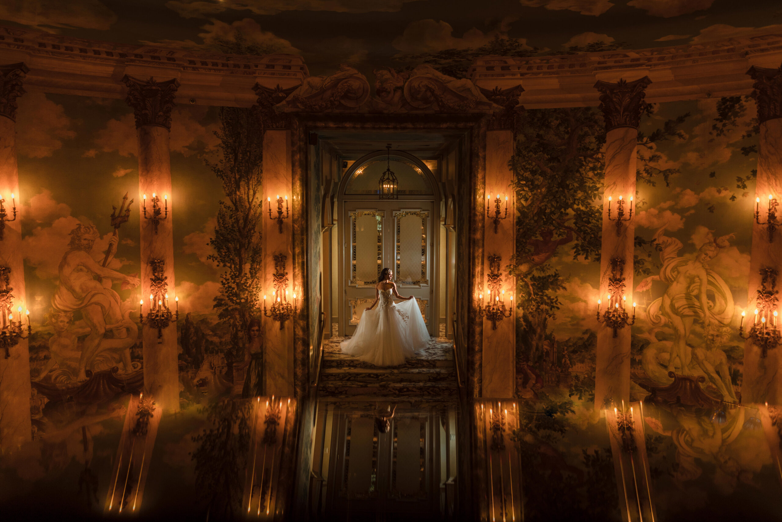 A bride in a white gown poses in a grand, ornate hall with murals and candlelit sconces. She stands facing away, bathed in warm light, with her reflection on the glossy floor captured perfectly by Boutique Wedding Photography in NYC.