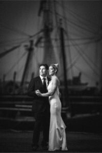 A man in a suit and a woman in a white dress stand together, holding hands and gazing into the distance, with a ship’s rigging blurred in the background—capturing the charm of Mystic CT wedding venues.