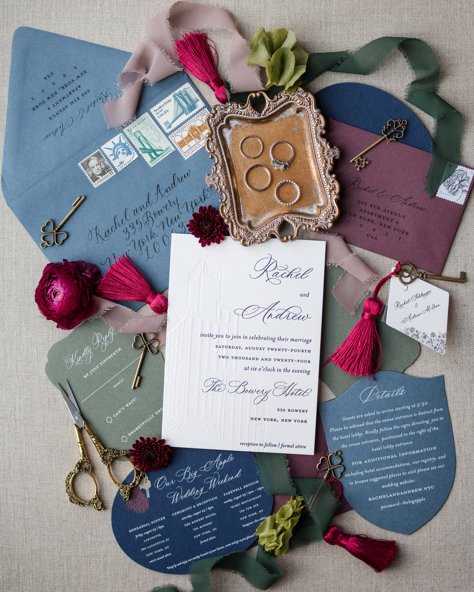 Flat lay image of wedding invitations in various shades of blue and maroon, surrounded by decorative keys, ribbons, scissors, flowers, and a bronze tray holding four coins—perfect props for capturing Bowery Hotel wedding photos.