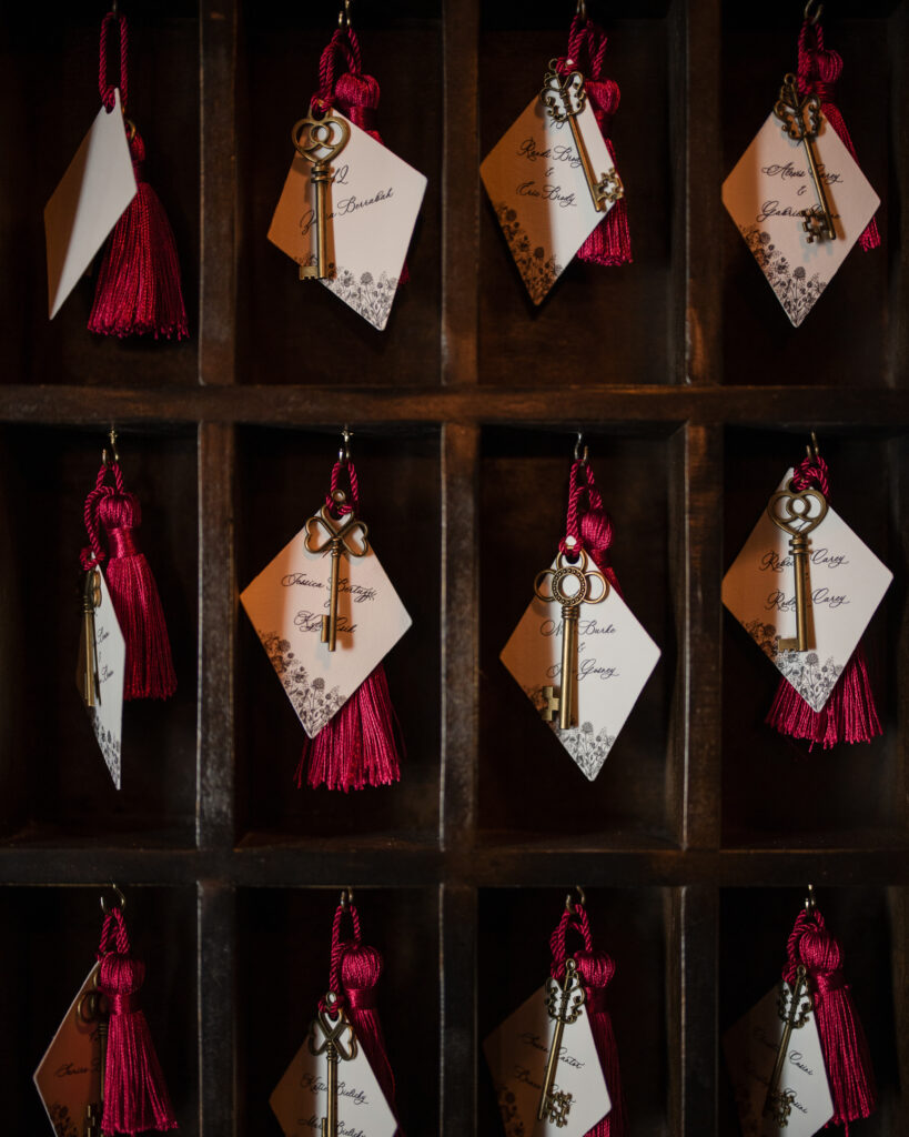 A wall-mounted wooden display at the Bowery Hotel wedding holds labeled brass keys with red tassels, each tag featuring names and table numbers, meticulously arranged in a grid pattern.