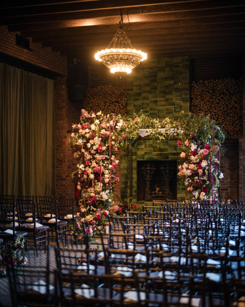 Indoor wedding venue with dark wooden chairs arranged in rows, defined by a green tiled fireplace. A floral arch with vibrant pink and white flowers stands beneath the overhead chandelier, set against rustic brick walls, perfect for capturing stunning Bowery Hotel wedding photos.