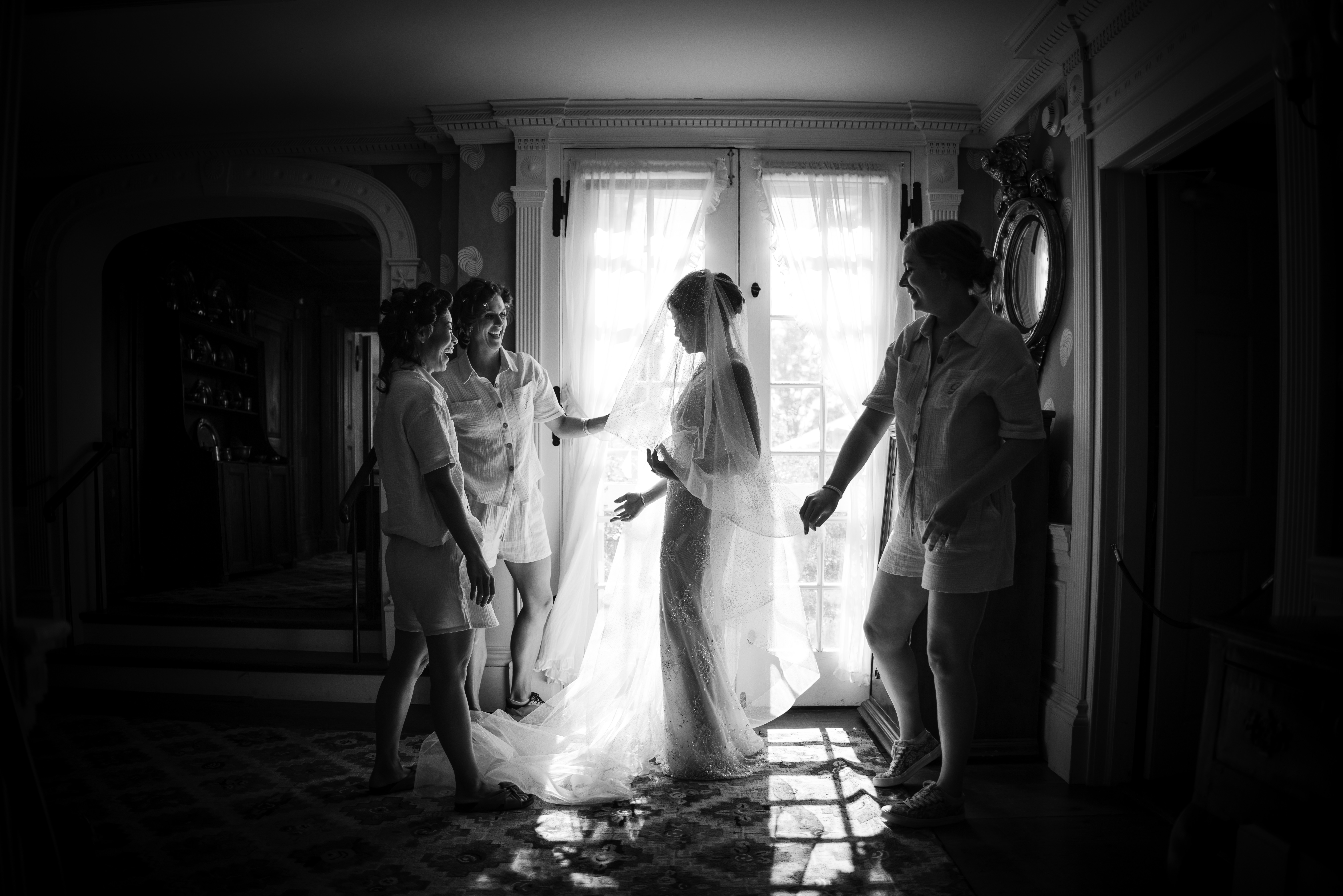 A bride stands illuminated by window light, wearing a veil and dress, surrounded by three people in casual attire, in a room with ornate decorations and a patterned floor—truly showcasing the elegance of boutique wedding photography in NYC.