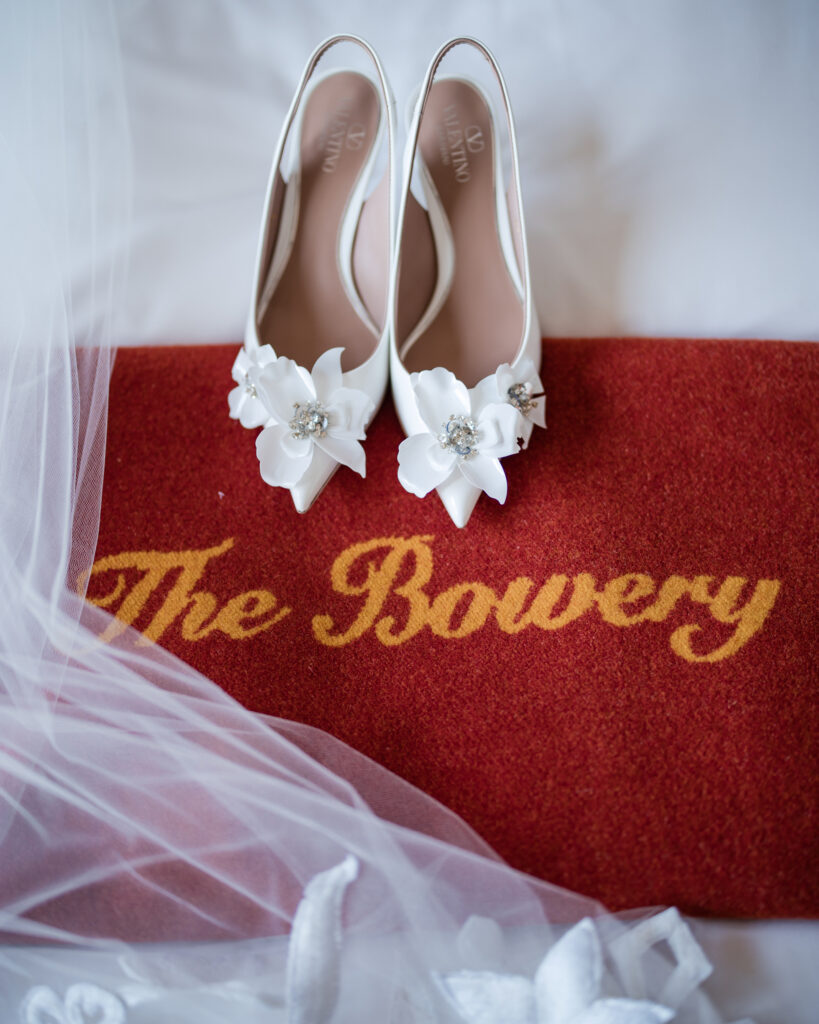 A pair of white floral-embellished high heels on a red mat with the words "The Bowery" in yellow, partially covered by white tulle fabric, capturing the essence of Bowery Hotel wedding photos.