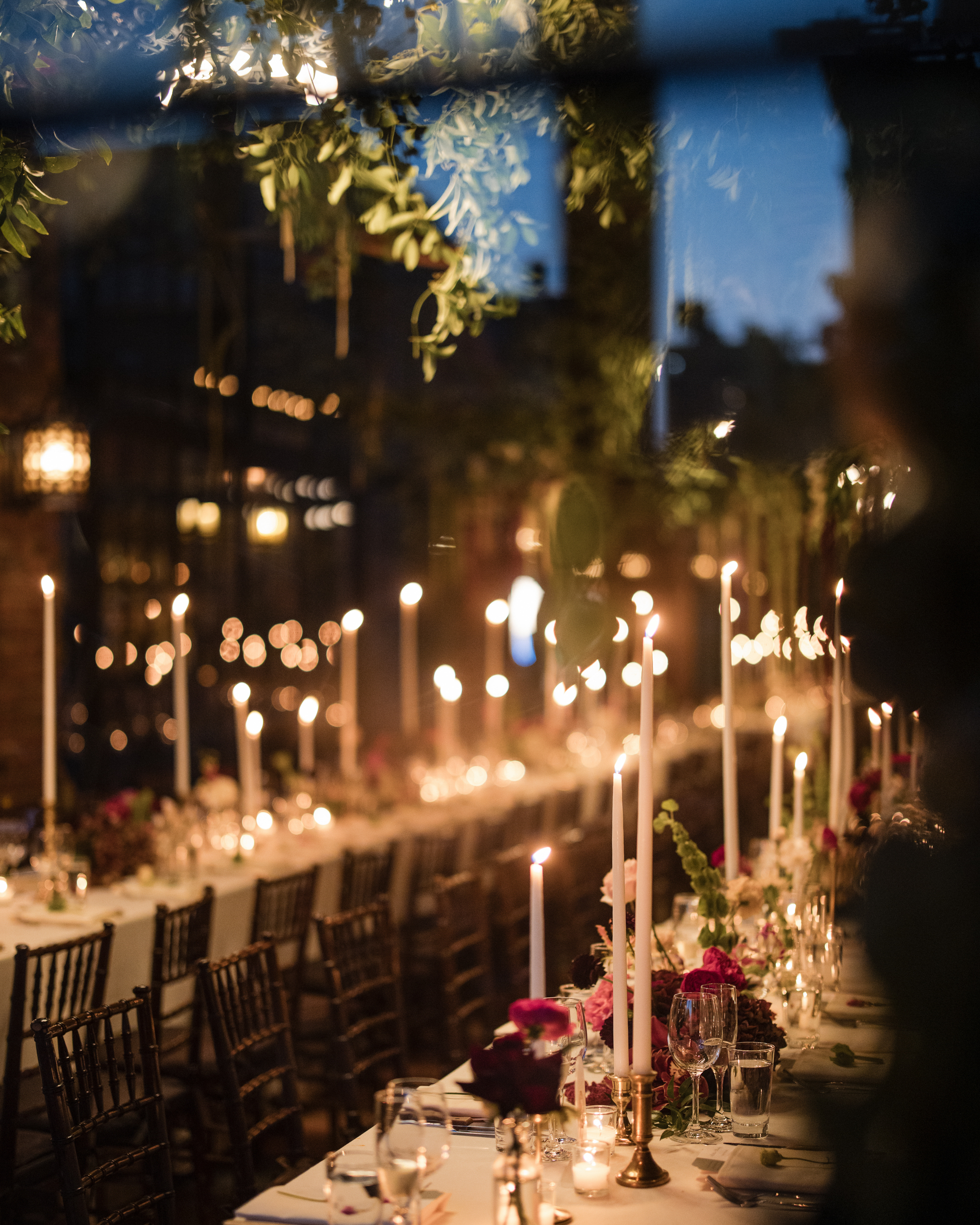 A long, elegantly set dining table with lit candles, floral centerpieces, and dark wooden chairs, reminiscent of a Bowery Hotel wedding photo, situated in a dimly lit, decorated outdoor or semi-outdoor setting.