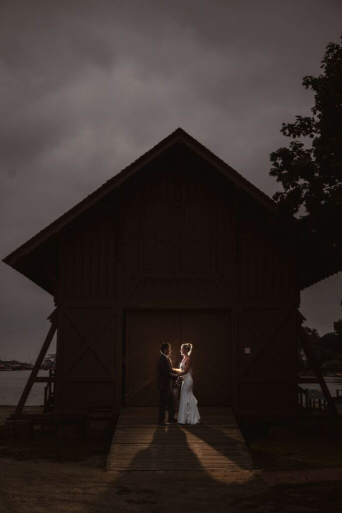 A couple stands in front of a dark, wooden structure, at Mystic CT wedding venues.