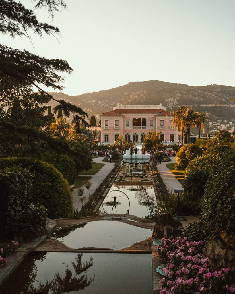 A pink villa with an elaborate garden featuring ponds and lush greenery, Villa Ephrussi de Rothschild offers a perfect setting for a wedding against the backdrop of hills under a clear sky.