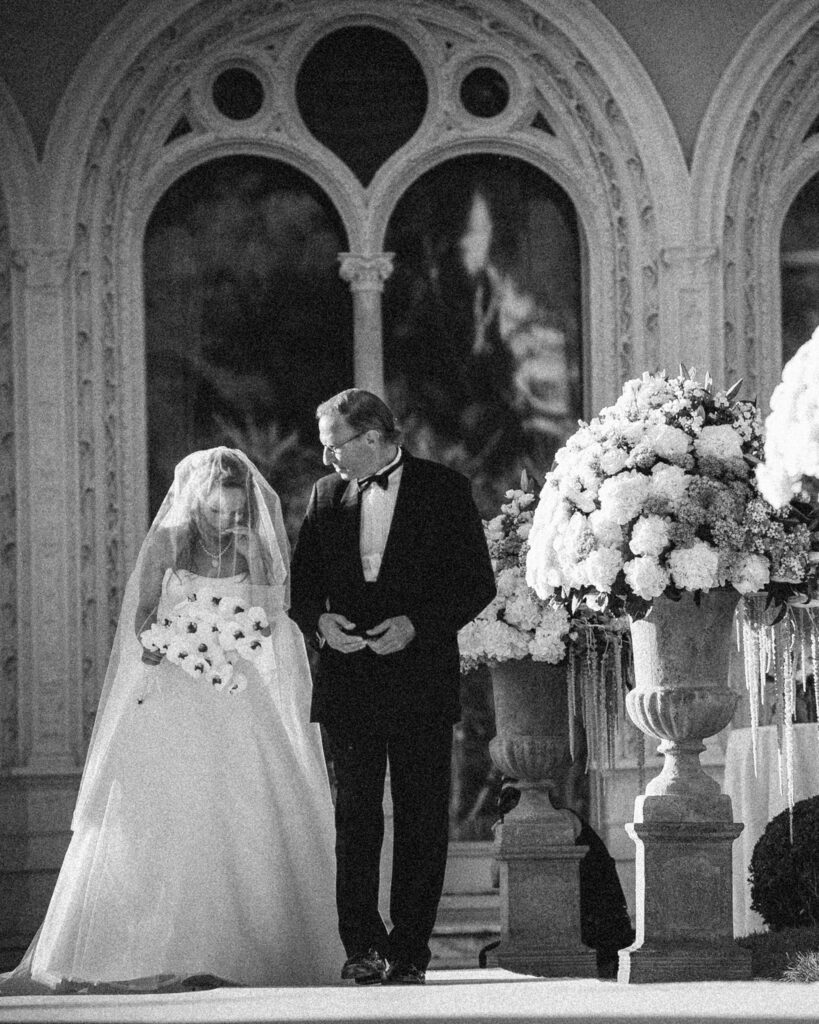 A bride and groom stand together in front of an ornate building with large arched windows, flanked by two large floral arrangements. The black-and-white scene evokes timeless elegance, reminiscent of a Villa Ephrussi de Rothschild wedding.