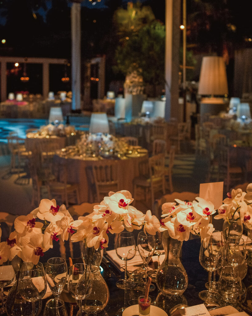 A dimly lit, elegantly set Villa Ephrussi de Rothschild wedding features round tables adorned with white orchids in vases, surrounded by chairs. In the background, more tables with similar decor are visible.