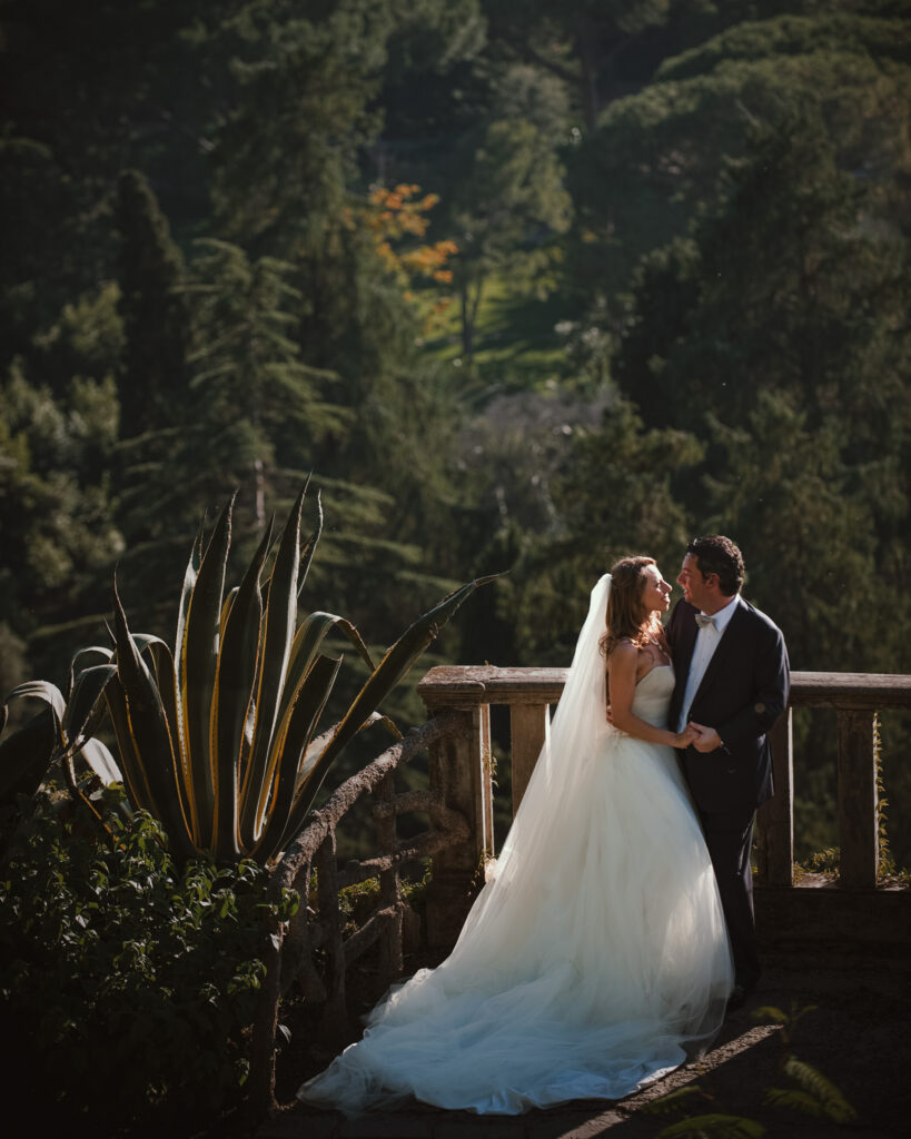 A bride and groom stand on a stone balcony surrounded by lush greenery, sharing an intimate moment in wedding attire. The bride wears a white dress with a veil, and the groom is in a dark suit, capturing the romance of their Villa Ephrussi de Rothschild wedding.