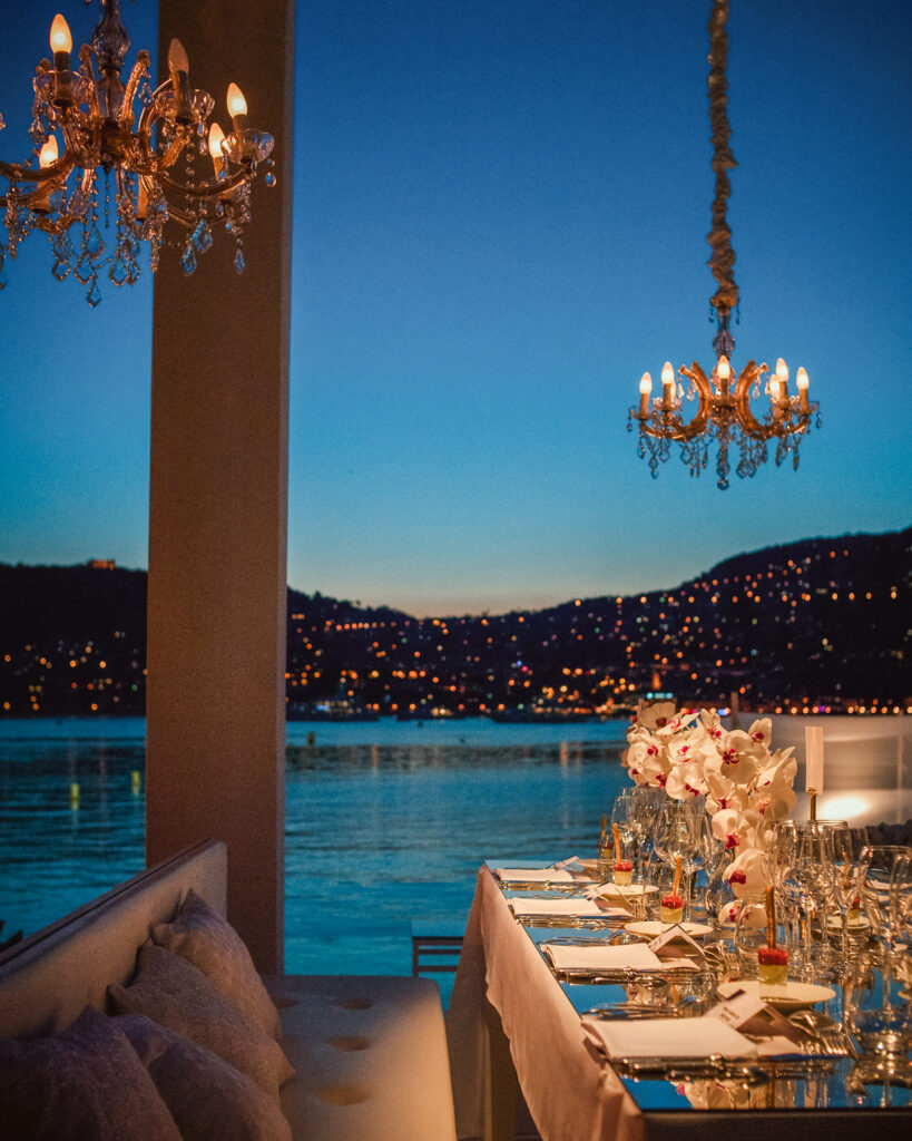 Elegant dining table setup with chandeliers overlooking a calm waterfront and the illuminated hills at dusk, reminiscent of a Villa Ephrussi de Rothschild wedding.