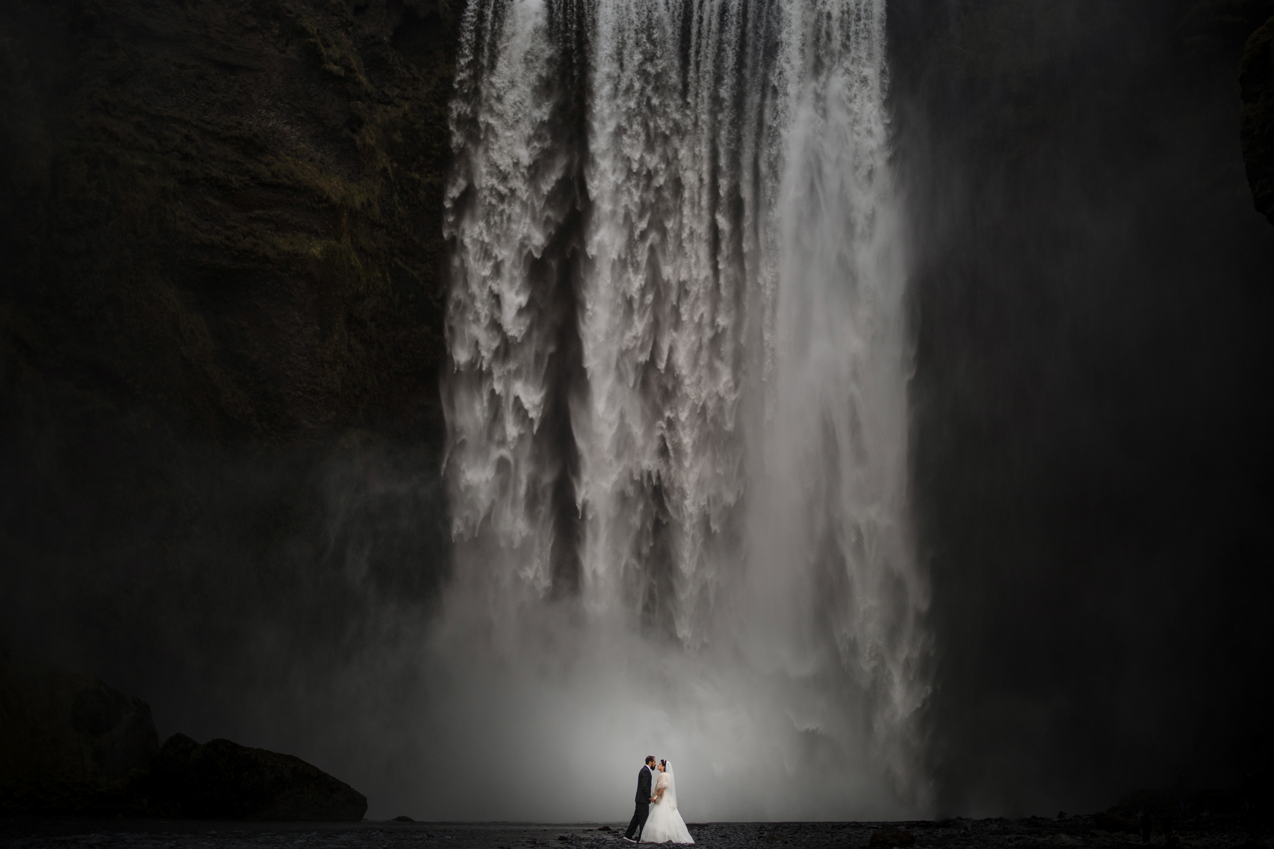 A bride and groom stand together in front of Iceland’s misty Skogafoss waterfall, surrounded by dark, rocky cliffs.
