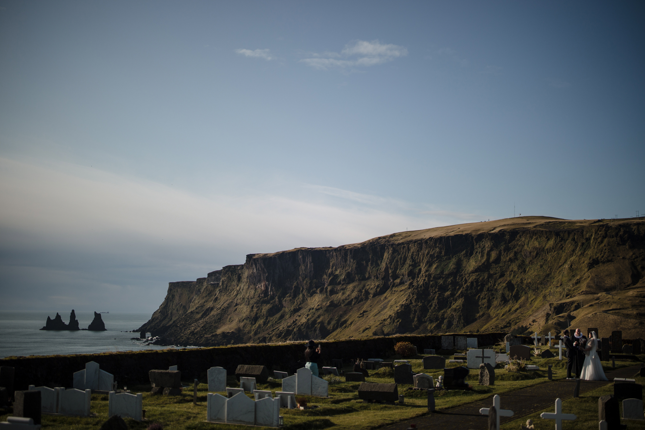 A bride and groom stand in a coastal cemetery with white headstones, overlooking cliffs and sea stacks under a clear sky—an unforgettable Iceland wedding.