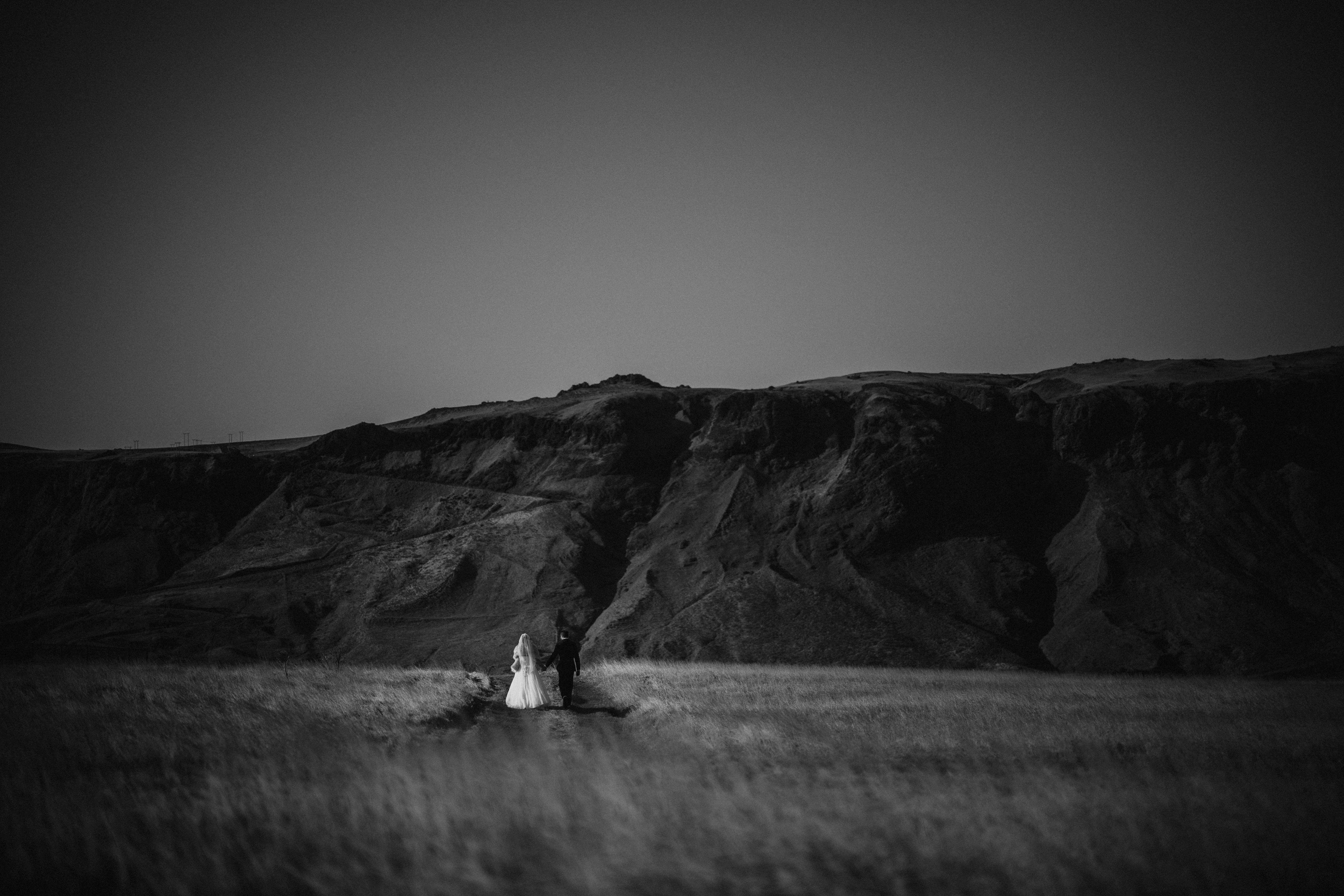 MASTER A bride and groom walk together across a grassy field towards rocky hills under a clear sky, in a black and white photograph in Vik at their Iceland destination wedding
