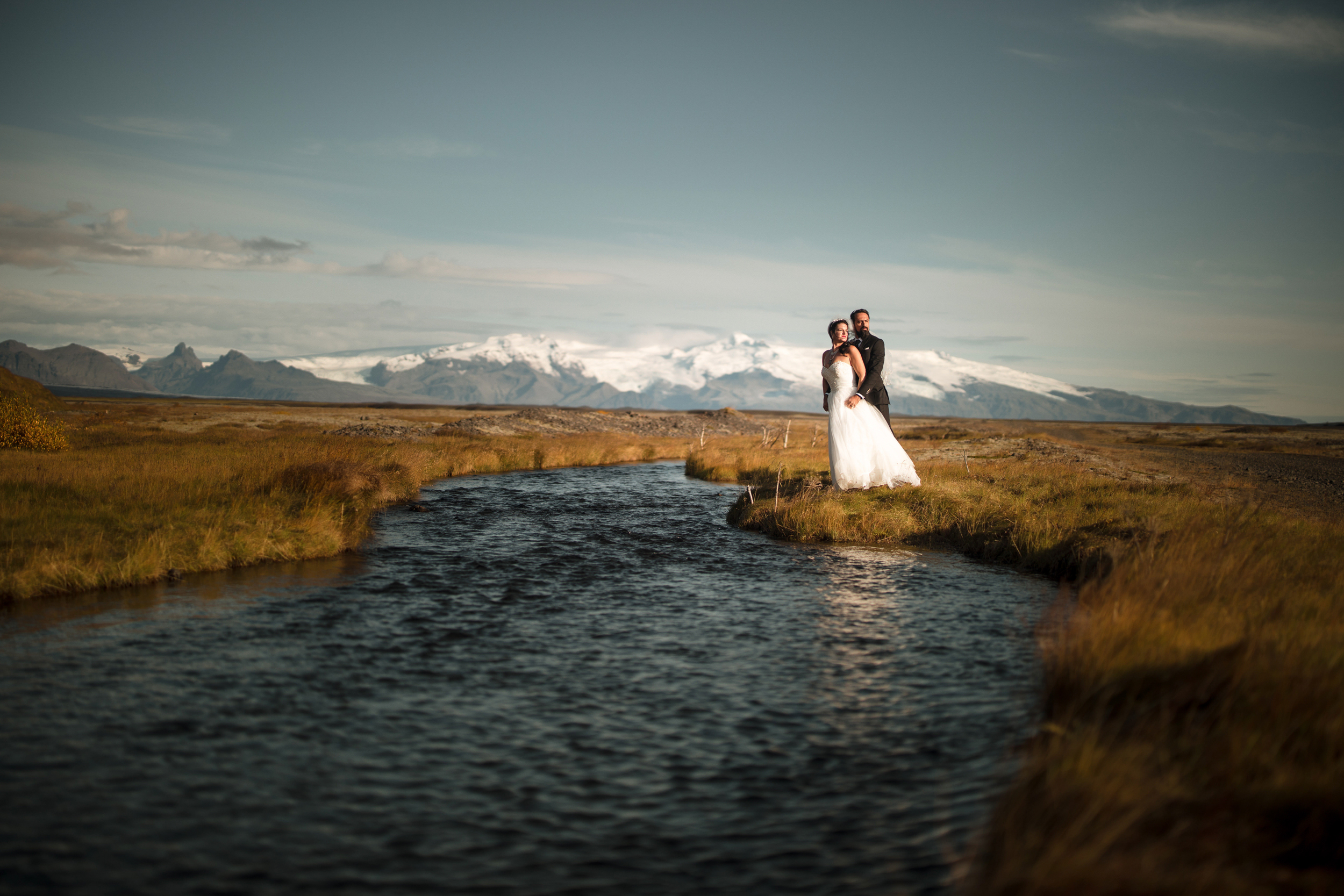 A couple in wedding attire stands beside a stream in a grassy field with mountains and snow in the background under a clear sky at an iceland destination wedding