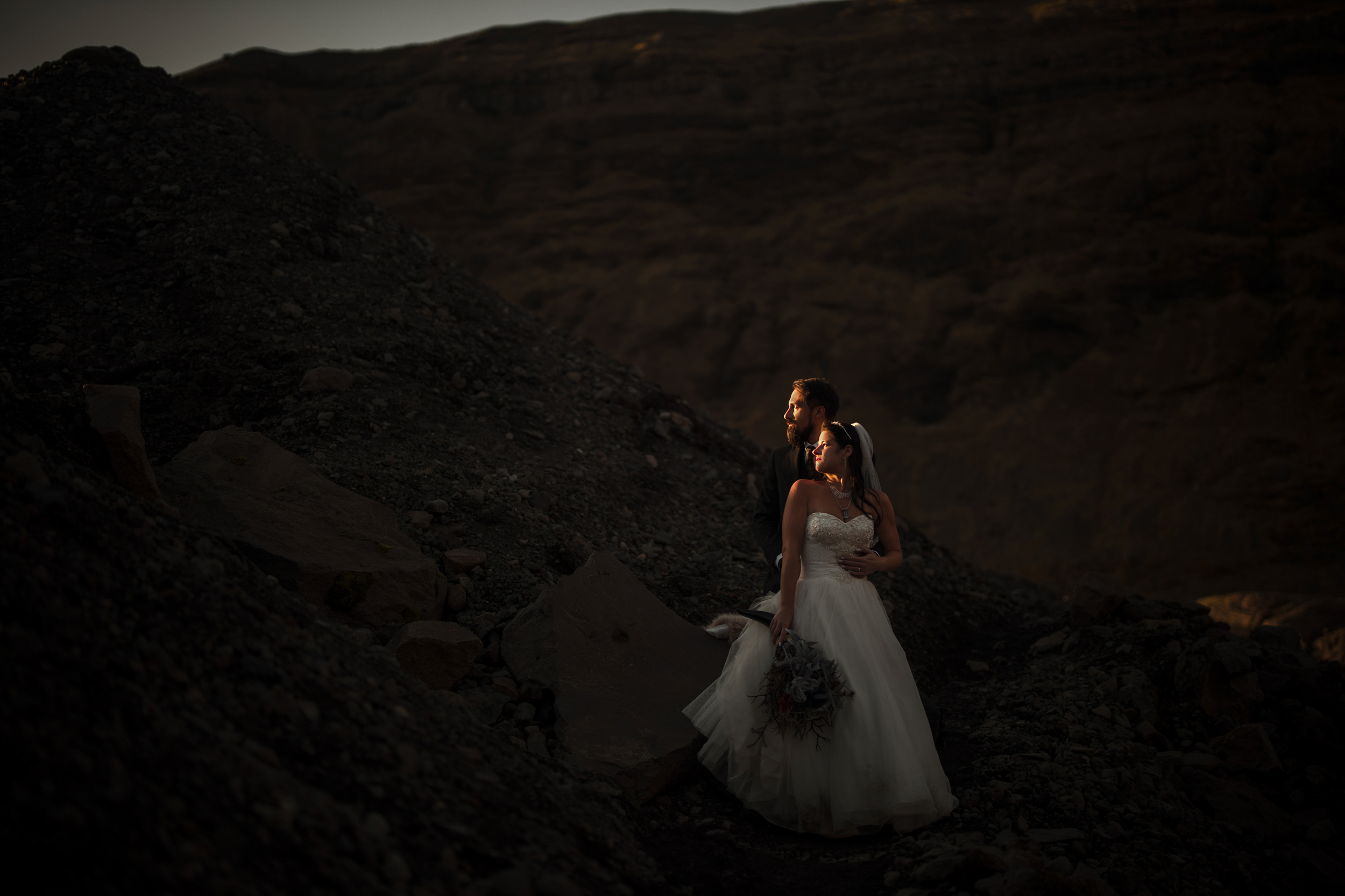 A couple in wedding attire stands on rocky terrain near Iceland’s Vik black sand beach, illuminated by soft light against a dark, mountainous background and dramatic basalt columns.