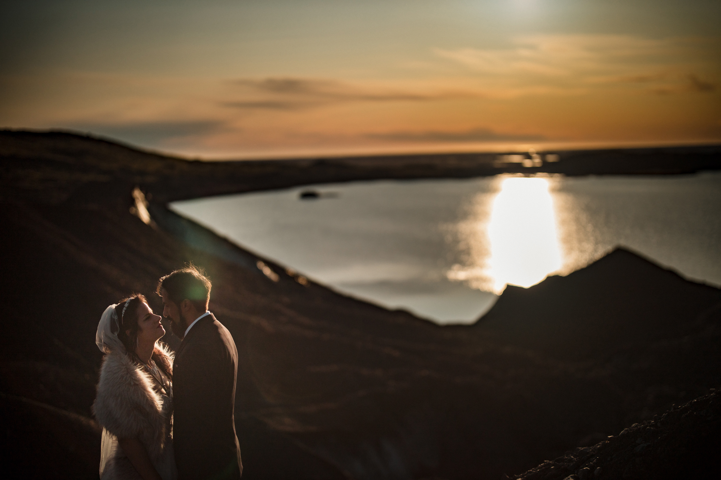 A couple stands close together by a lake at sunset, with hills silhouetted against the bright sky and water, evoking the romance of an Iceland wedding near a glacier.