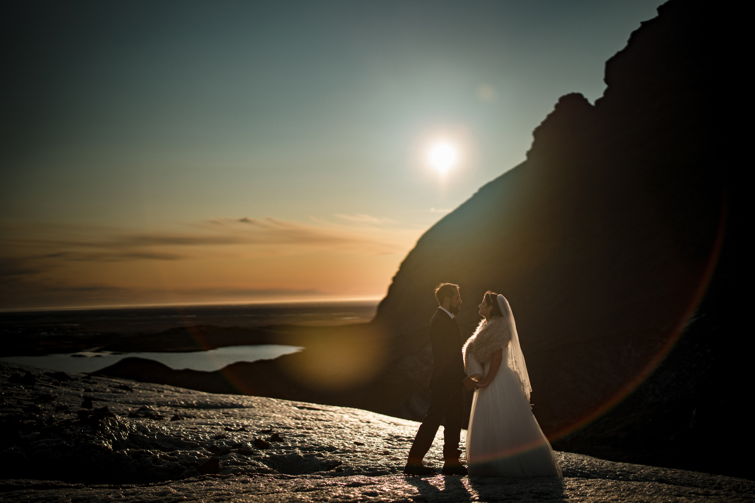 A bride and groom stand together on rocky terrain at sunset, silhouetted against the sun with a mountain and body of water in the background, capturing the wild beauty of an Iceland wedding.