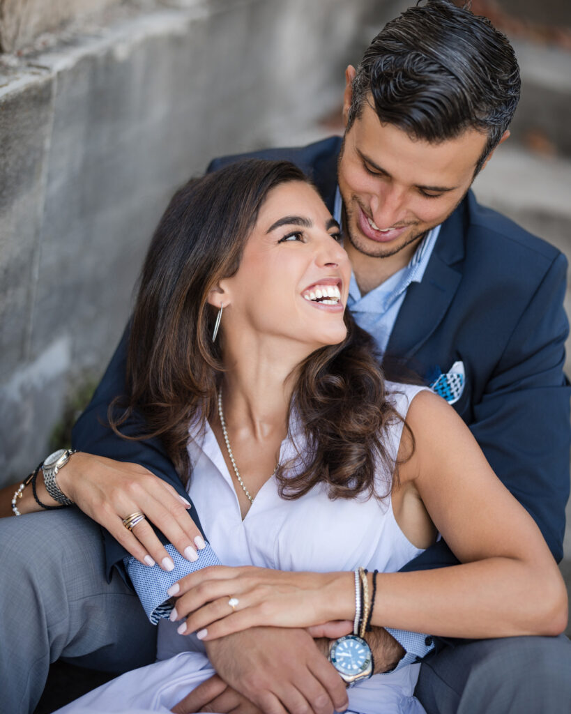 A couple seated outdoors at Untermyer Gardens, embracing and smiling at each other. The man is wearing a suit, and the woman is in a white outfit with jewelry, capturing the perfect engagement moment.