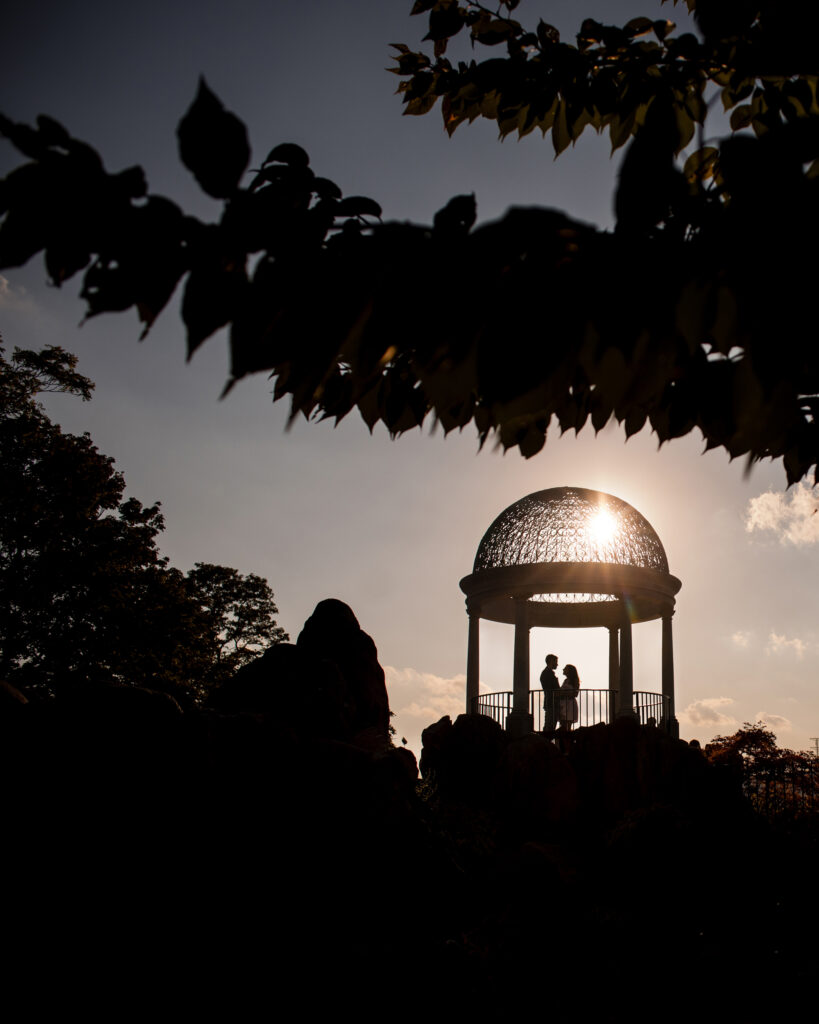 A silhouetted couple stands inside a gazebo with a dome, partially obscured by tree leaves, against a sunset sky at Untermyer Gardens, capturing the essence of an enchanting engagement.