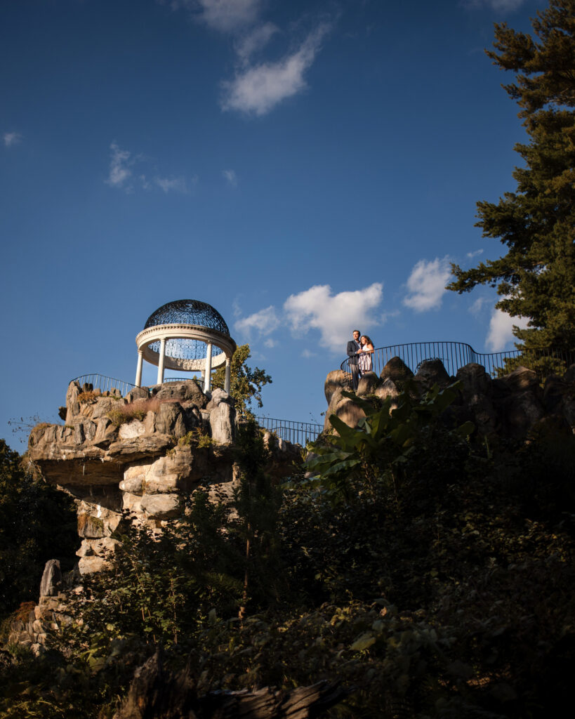 A couple stands on a rocky outcrop with a metal gazebo under a blue sky with scattered clouds, capturing the magic of an Untermyer Gardens engagement. Trees weave through the background, adding to the enchanting scenery.