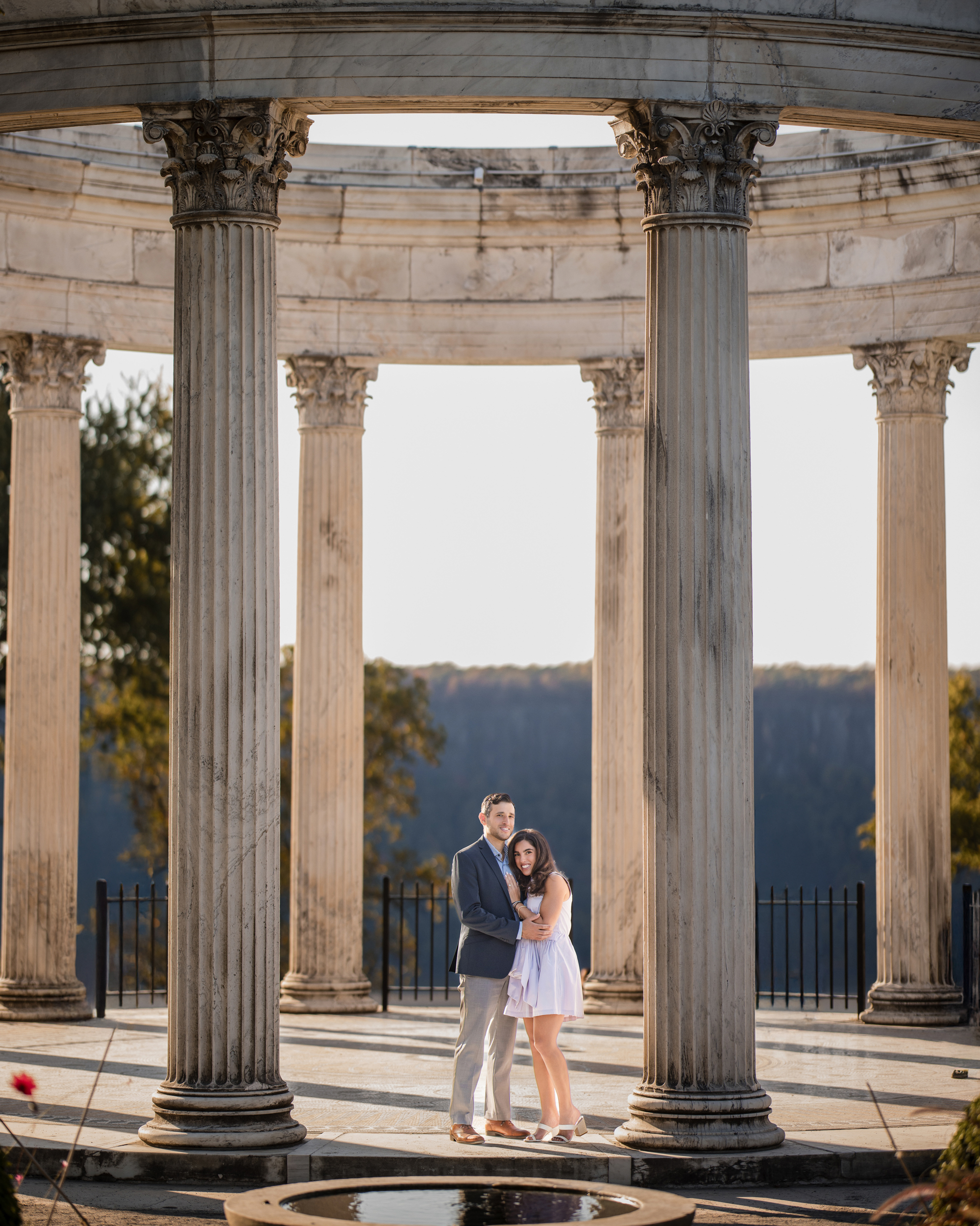A couple stands close together under the circular colonnade of Untermyer Gardens, embraced by tall pillars. Trees and a scenic view enhance the enchanting backdrop of their engagement moment.