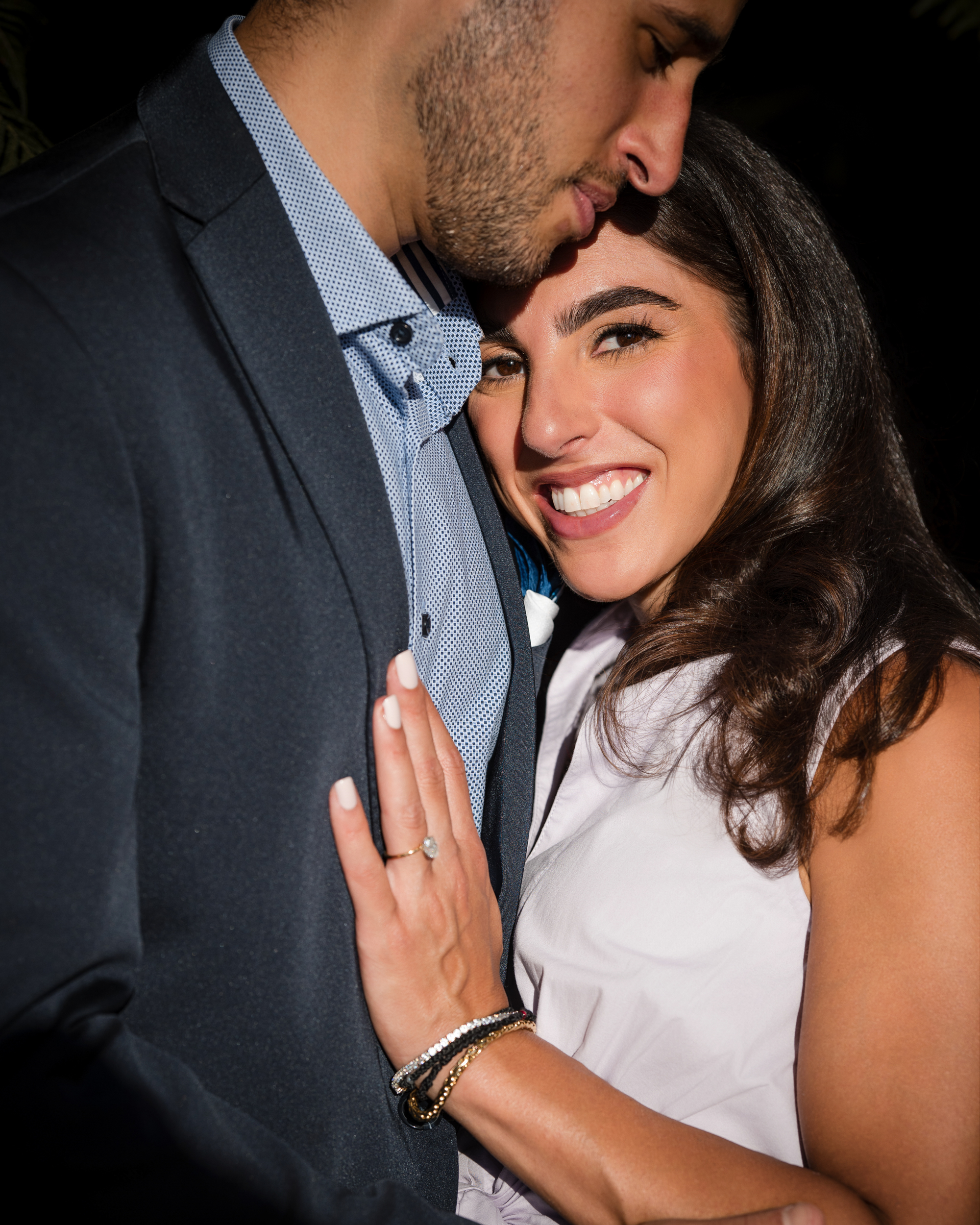 A woman beams as she embraces a man at Untermyer Gardens, proudly displaying an engagement ring on her finger. The man, dressed in a suit, mirrors her joy while the woman shines in her sleeveless top.