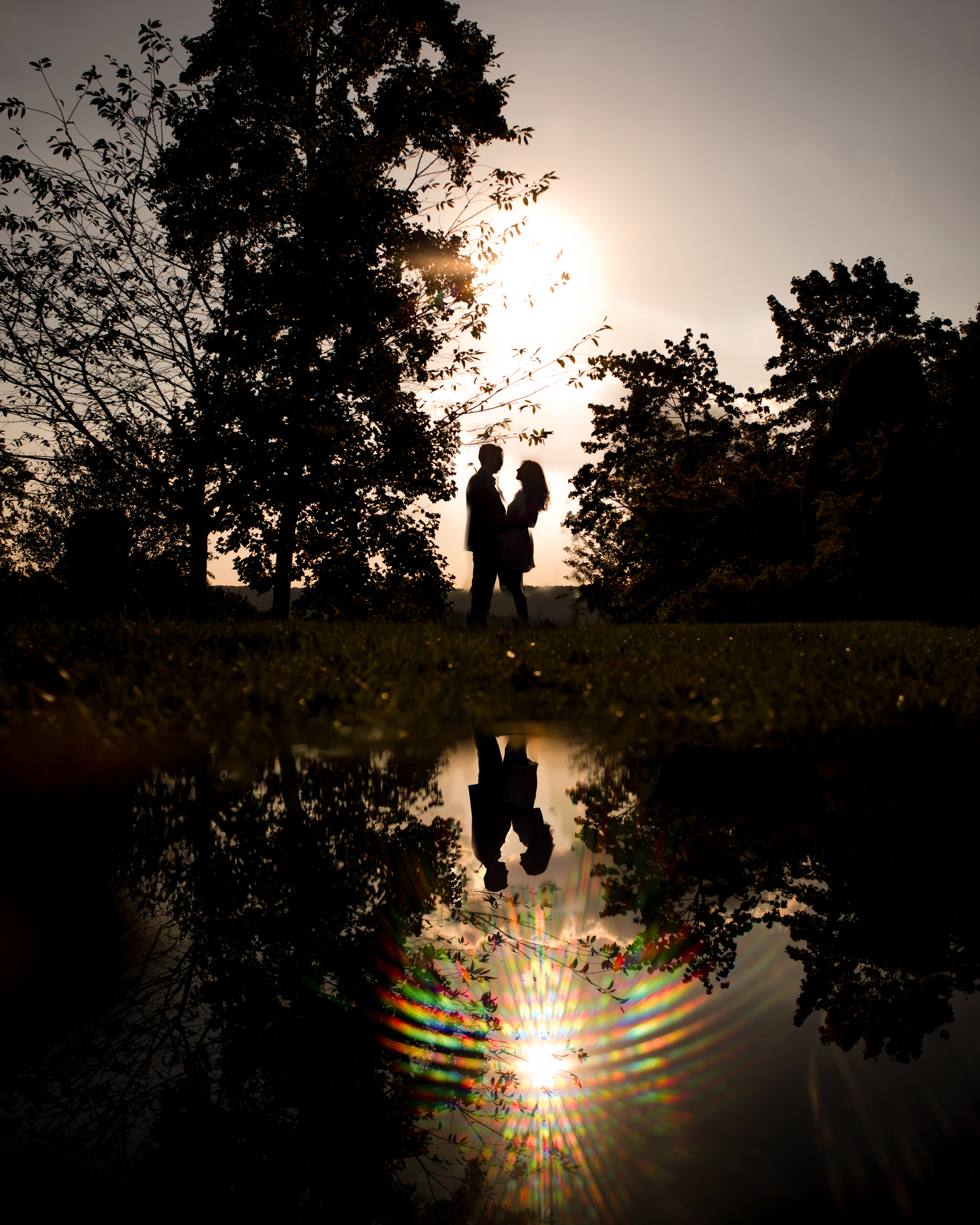 Silhouetted couple stands by a tree at sunset in Untermyer Gardens, their reflection shimmering in the pond below as light creates a spectrum effect.