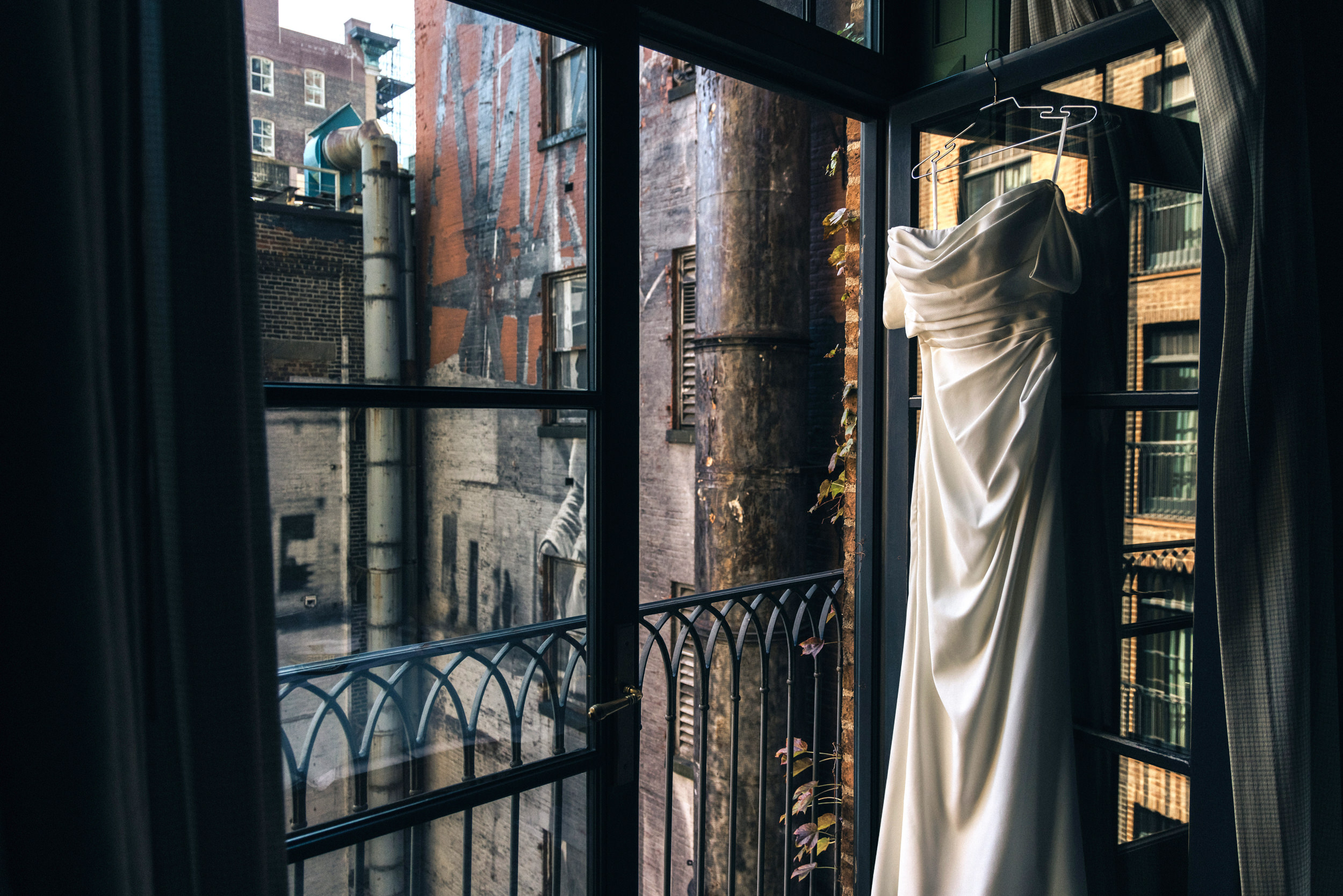 A white dress hangs on a hanger by a window at the Greenwich Hotel, offering an urban view with brick buildings and metal structures—a perfect setting for a wedding.