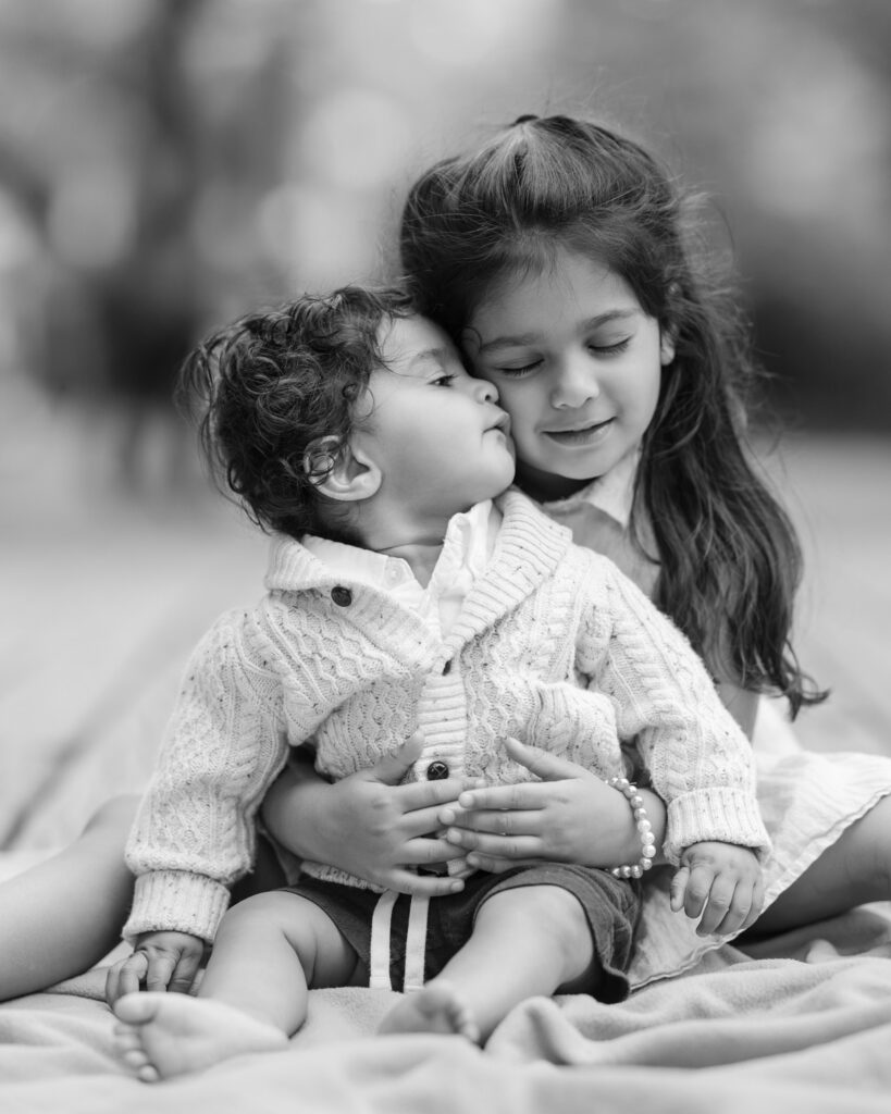 A black and white photo captures a young girl holding her baby sibling on her lap during a visit to Central Park. Both are wearing cozy sweaters. The girl smiles warmly as the baby leans in affectionately, preserving a cherished family moment in an iconic setting.