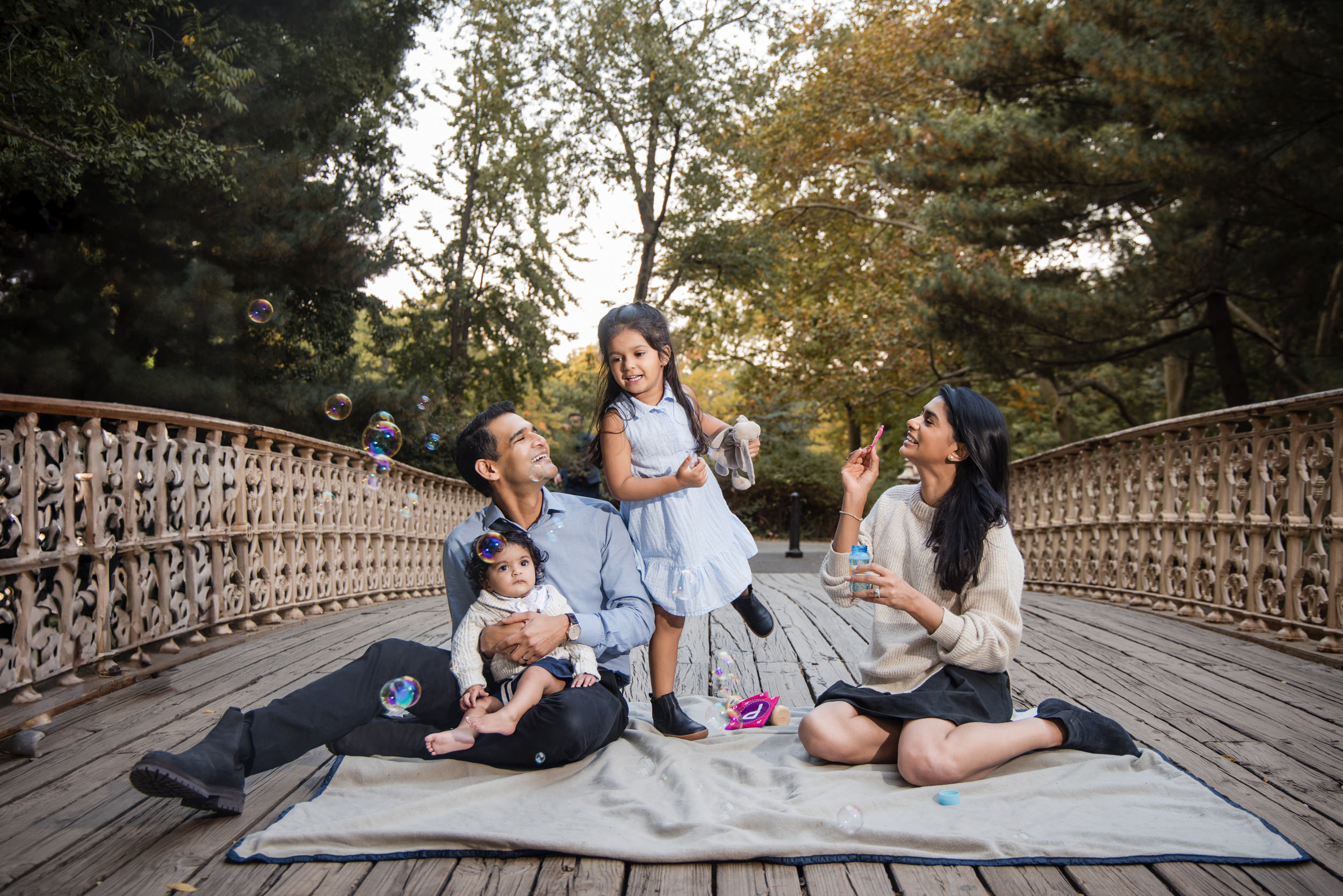 A family of four sits on a blanket on a wooden bridge in Central Park, surrounded by trees. A woman blows bubbles as a man holds a baby, and a young girl stands between them, smiling.