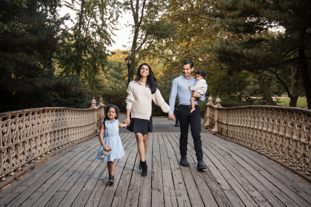 A family of four strolls across a wooden bridge in Central Park. The parents hold hands with a young girl, while the father carries a toddler. Lush trees envelop them, capturing an idyllic family moment perfect for photos.