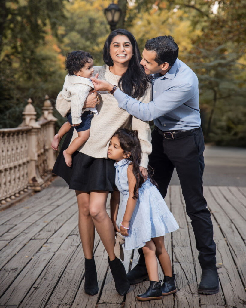 A family with two children stands on a wooden bridge in Central Park, smiling. The father holds one child, and the mother and daughter are standing close together. Lush trees create a picturesque backdrop for this charming family photo.