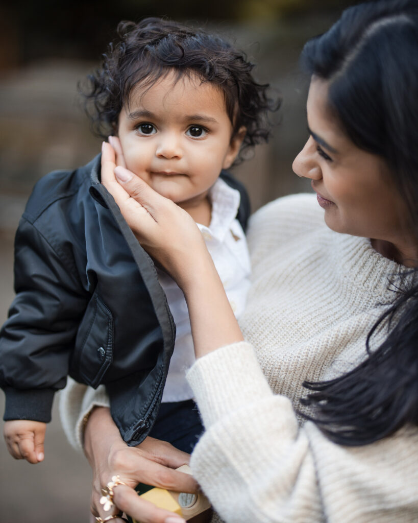 In Central Park, a woman in a white sweater holds a toddler in a black jacket, gently touching the child's cheek—a tender moment captured in family photos.