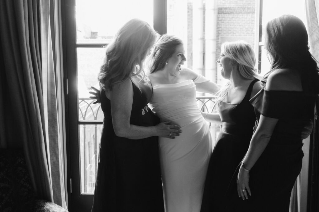 Four women in formal dresses stand by a window at the Greenwich Hotel, smiling and talking. One woman is in a bridal gown, capturing the elegance of a wedding moment. The room is softly lit, enhancing the intimate atmosphere.