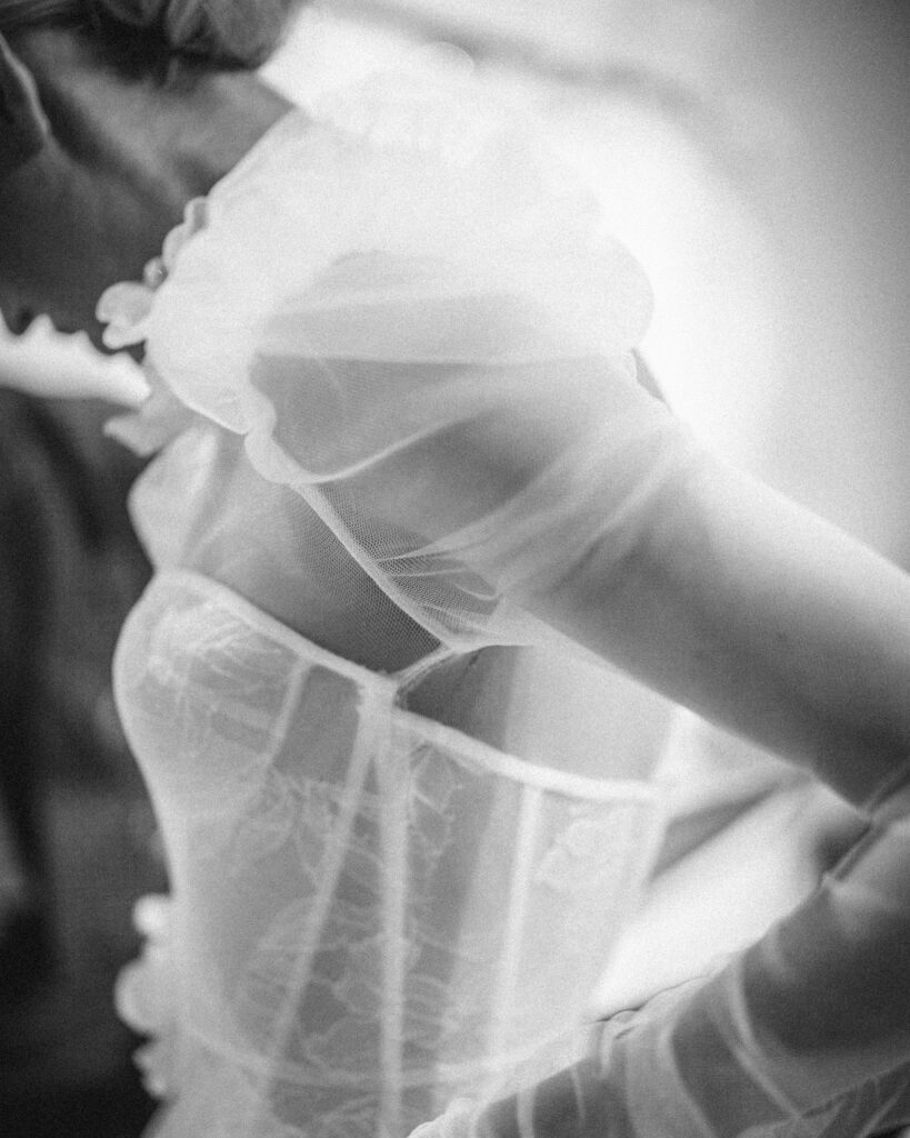 Close-up black and white image of a person wearing a sheer, delicate garment with puffy sleeves from the Pnina Tornai AURA Collection. The focus is on the fabric's intricate texture and design, capturing an ethereal elegance.