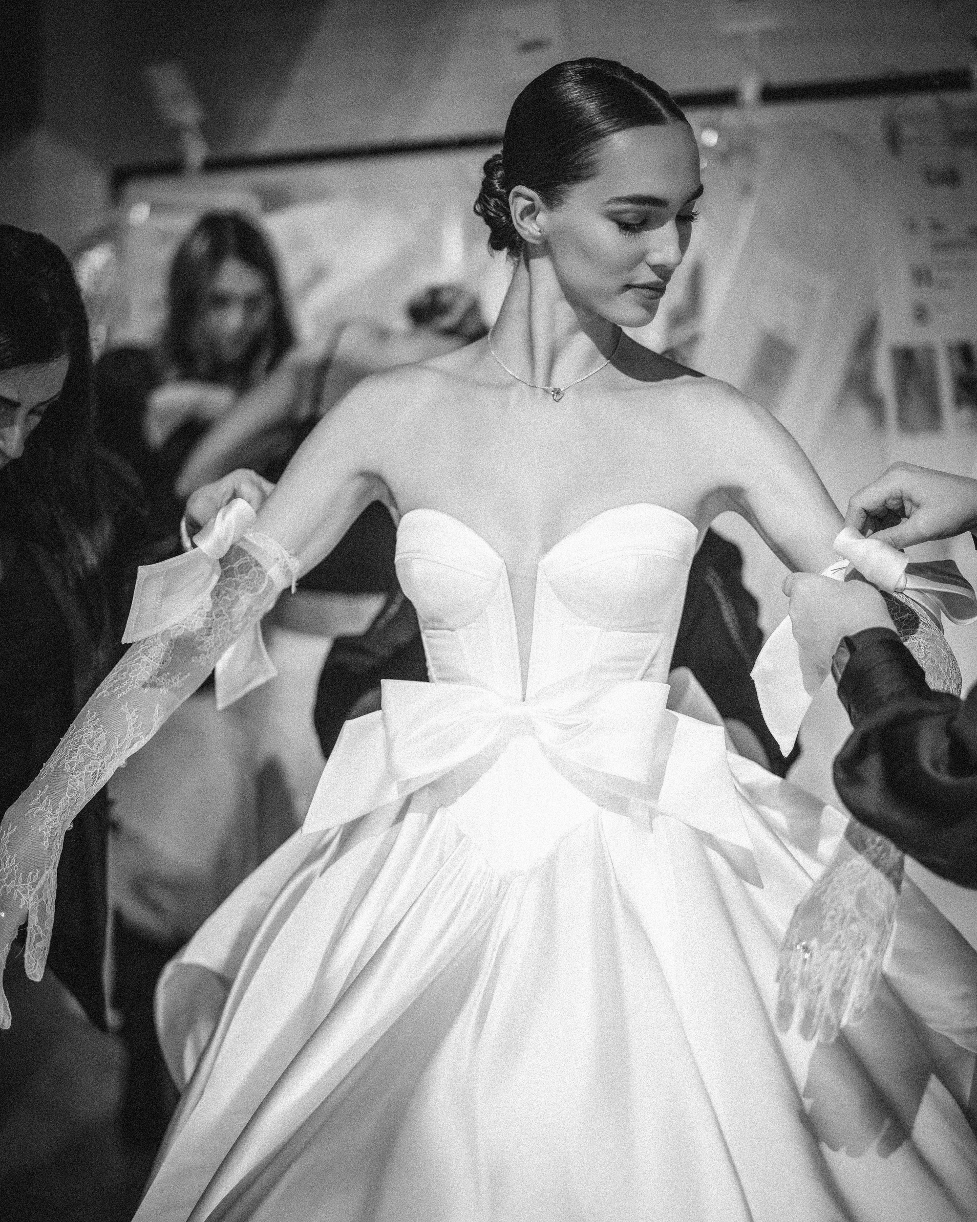 A woman in a white gown from the Pnina Tornai AURA Collection, adorned with bows and lace gloves, is gracefully attended by others in the background.