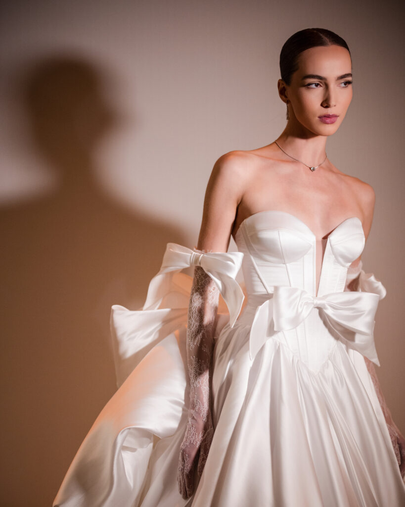 A model poses in an elegant white gown from the Pnina Tornai AURA Collection, complete with a bow, long gloves, and a necklace, against a neutral background.