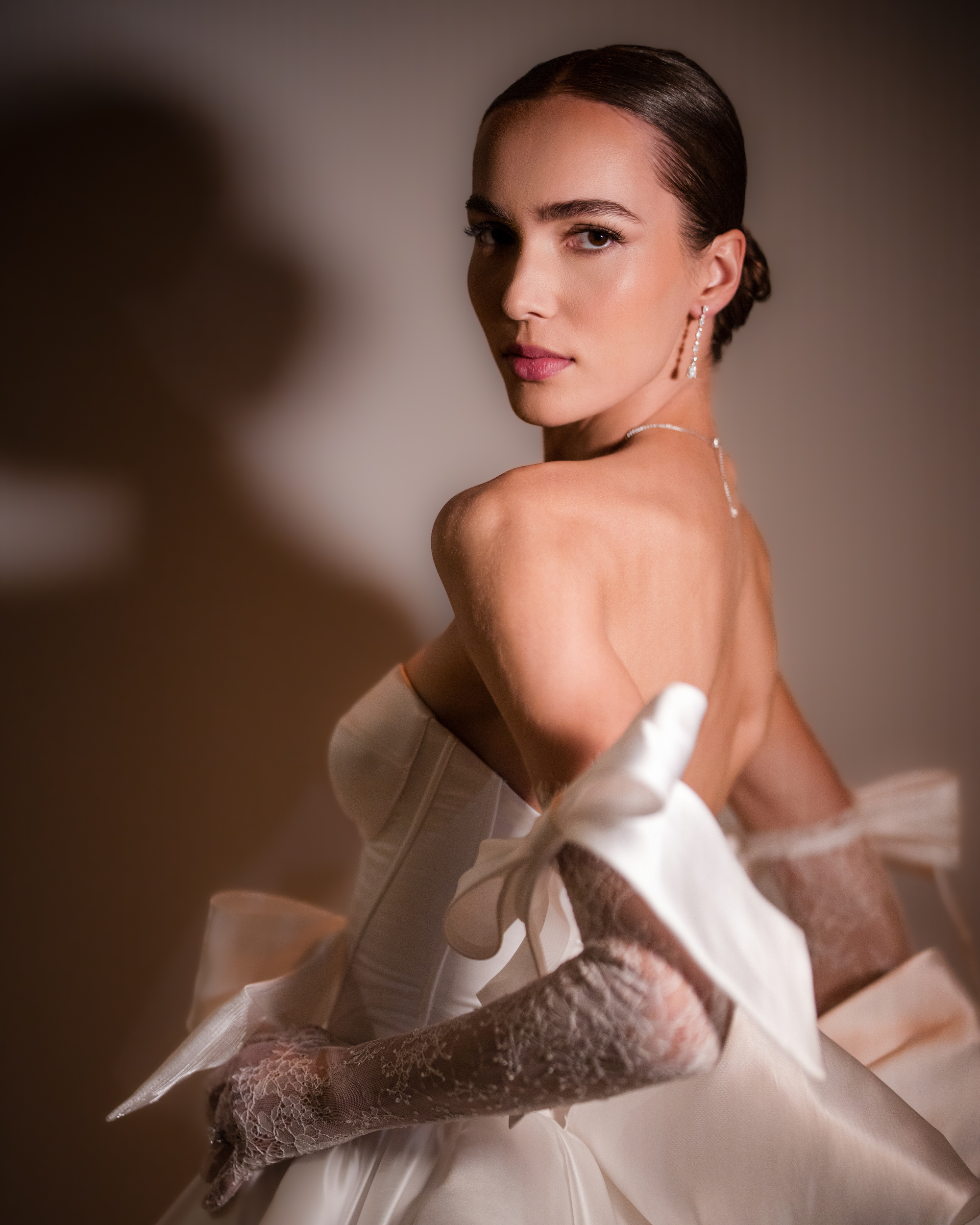 Person in an elegant white strapless dress from the Pnina Tornai AURA Collection, with lace gloves, looking over their shoulder against a soft-lit background.