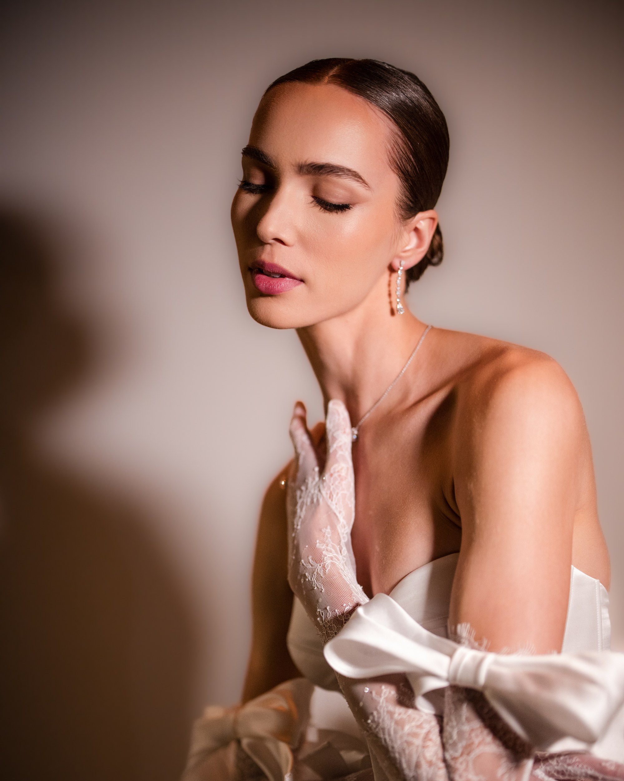 A woman in a strapless dress from the Pnina Tornai AURA Collection, adorned with lace gloves and earrings, poses with eyes closed against a neutral background.