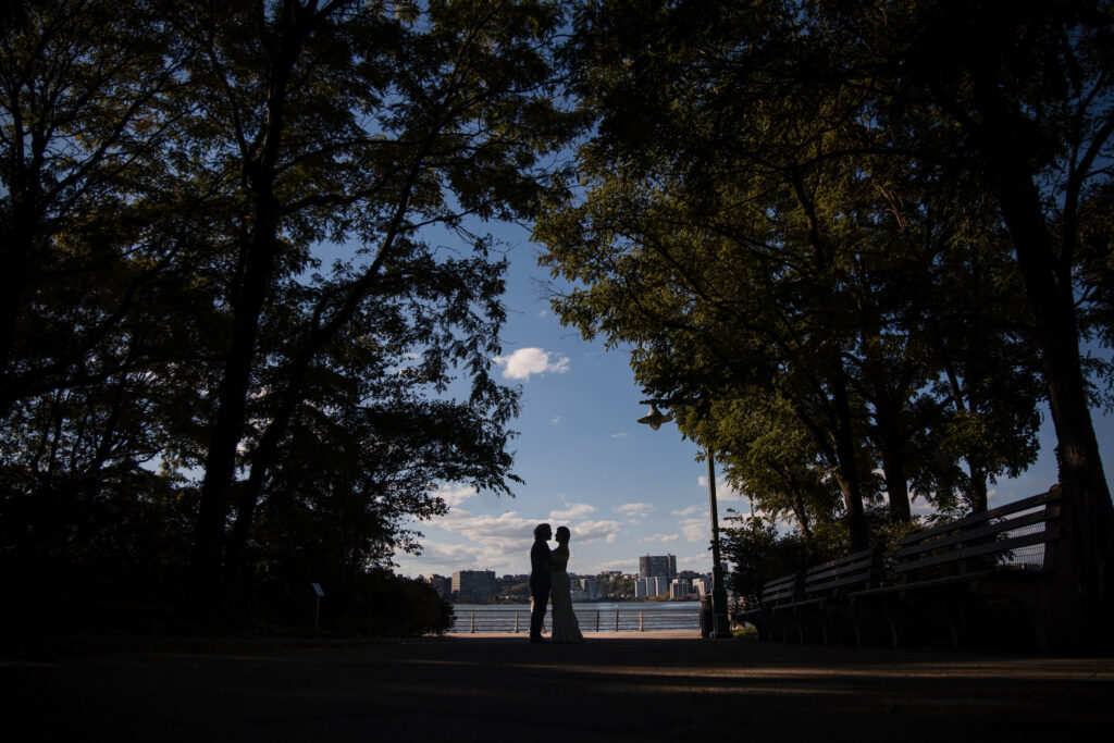 Silhouetted couple under trees by a river, with the cityscape in the background and benches along the path, captures a moment as enchanting as a Greenwich Hotel wedding. 